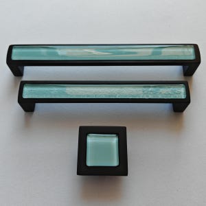May include: Set of three cabinet hardware pieces: two rectangular pulls and one square knob. The hardware features a black finish with a light blue, textured glass insert. The pulls are different sizes, and the knob is square.