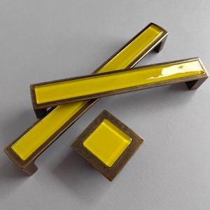 Può includere: Un set di ferramenta per mobili con un inserto rettangolare giallo brillante all'interno di una cornice color bronzo. Il set include due maniglie lunghe e rettangolari e un pomello quadrato. Per mobili e decorazioni per la casa.