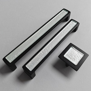 Op de afbeelding: Een set zwarte meubelbeslag, bestaande uit twee rechthoekige handgrepen en een vierkante knop. Elk stuk heeft een getextureerd, zilverkleurig middenpaneel. Voor meubels en kasten.