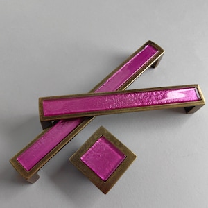 Może przedstawiać: Zestaw trzech różowych szklanych gałek do szafek z mosiężnym wykończeniem. Gałki są prostokątne i kwadratowe.