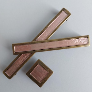 Op de afbeelding: Drie kastgrepen met een geborsteld messing afwerking en een roze glazen inzet. De grepen zijn rechthoekig en hebben afgeronde randen. De kleinere greep is vierkant.