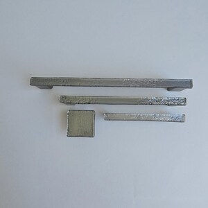 May include: A collection of silver-colored cabinet hardware, including two rectangular handles, a square knob, and a smaller rectangular pull. The hardware has a textured, hammered appearance, suggesting a handcrafted or rustic design. The pieces are arranged on a white surface.