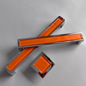 Op de afbeelding: Een set kastbeslag met een feloranje rechthoekig middenstuk, omlijst door een gepolijste chromen lijst. De set bevat twee lange handgrepen en een vierkante knop, allemaal met een modern design.