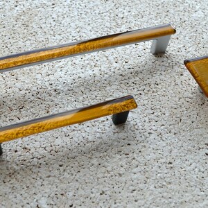 Pode incluir: Um conjunto de ferragens para armários com uma pega longa e retangular com postes prateados, uma pega mais pequena com postes pretos e um puxador quadrado. A ferragem tem um centro transparente de cor âmbar.