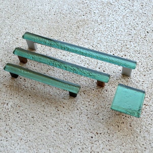 May include: A collection of four aqua-coloured glass cabinet pulls and a knob. The rectangular pulls have a textured surface and are mounted on silver, brown, and black hardware. The square knob has a matching aqua colour and texture.