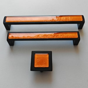 Può includere: Set di tre pezzi di ferramenta per mobili: due maniglie rettangolari e un pomello quadrato. La ferramenta presenta una finitura nera opaca con un inserto in vetro arancione strutturato. Le maniglie sono lunghe circa 15 cm.