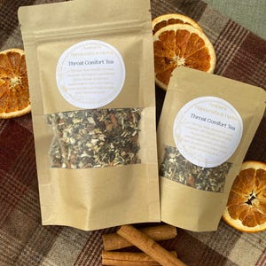 May include: Two kraft paper pouches of Throat Comfort Tea with a clear window, surrounded by dried orange slices and cinnamon sticks. The tea pouches have a white label with text. The tea blend includes ingredients like slippery elm and wild cherry.