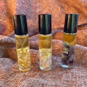 May include: Three small glass bottles with black roll-on tops filled with a clear liquid and small pieces of crystals. The bottles are on a brown fabric surface.