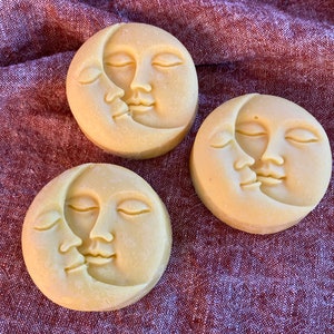 Pode incluir: Três sabonetes redondos amarelo pálido com um design de lua crescente e rosto. O rosto da lua está beijando um rosto menor. Os sabonetes estão em uma superfície texturizada marrom avermelhada.