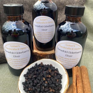 Op de afbeelding: Drie flessen vlierbessenelixer met zwarte doppen en etiketten met de tekst "Amber's Handcrafts & Herbs Threefold Elderberry Elixir". Een kleine kom met gedroogde vlierbessen en kaneelstokjes is ook zichtbaar.