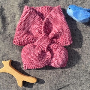 May include: A hand-knitted pink scarf with a twisted design. A small blue felt bird and a wooden fox-shaped toy are placed near the scarf on a gray surface. The scarf is made of a soft, textured yarn.