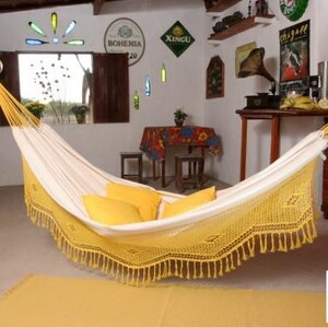 Pode incluir: Uma rede creme com franjas amarelas e almofadas amarelas est&aacute; pendurada em uma sala. A rede est&aacute; suspensa no teto. A sala tem uma decora&ccedil;&atilde;o r&uacute;stica com m&oacute;veis de madeira e objetos decorativos.