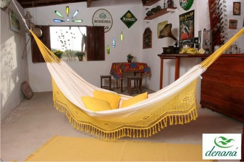 Pode incluir: Uma rede creme com franjas amarelas e almofadas amarelas est&aacute; pendurada em uma sala. A rede est&aacute; suspensa no teto. A sala tem uma decora&ccedil;&atilde;o r&uacute;stica com m&oacute;veis de madeira e objetos decorativos.