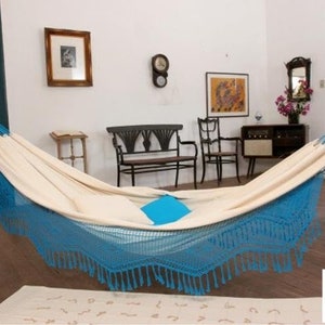 Pode incluir: Uma rede creme com franjas e detalhes azuis est&aacute; pendurada em uma sala com piso de madeira. A rede est&aacute; suspensa entre dois suportes azuis. Uma almofada quadrada azul est&aacute; dentro da rede.