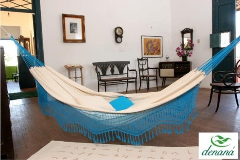 Pode incluir: Uma rede creme com franjas e detalhes azuis est&aacute; pendurada em uma sala com piso de madeira. A rede est&aacute; suspensa entre dois suportes azuis. Uma almofada quadrada azul est&aacute; dentro da rede.