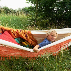 May include: A child is seen relaxing in a hammock. The hammock is white with orange netting and fringe. It is filled with colourful pillows and a patterned blanket. The scene is set in a grassy area with trees in the background.
