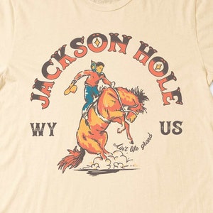 May include: A beige t-shirt with a vintage-style graphic of a cowboy riding a bucking horse. The graphic says "Jackson Hole WY US" and "Live like the West".