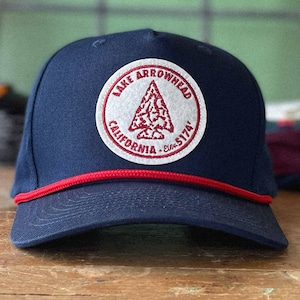 May include: A navy blue baseball cap with a red brim and a circular patch. The patch features the words "Lake Arrowhead California" around a red and white arrowhead design. The elevation is noted as 5174'.