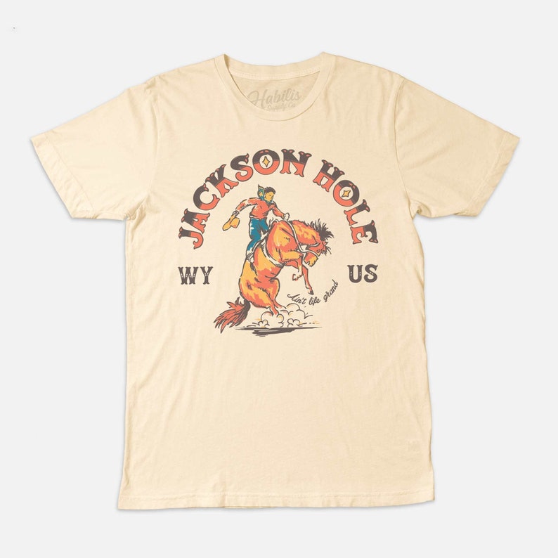 May include: A beige t-shirt with a vintage-style graphic of a cowboy riding a bucking horse. The graphic says "Jackson Hole WY US" and "Live like the West".