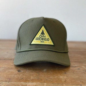 Könnte beinhalten: Olivgrüne Baseballkappe mit strukturierter Krone und gebogenem Schirm. Die Kappe hat einen gelben Dreieckaufnäher mit dem Text "Lake Arrowhead 1921" und einer Baumgrafik. Die Kappe liegt auf einer Holzoberfläche.
