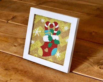 Puede incluir: Una obra de arte enmarcada con un calcetín navideño. El calcetín es rojo con ribetes blancos y está lleno de regalos y bastones de caramelo. El fondo es dorado con copos de nieve blancos. El marco es blanco.