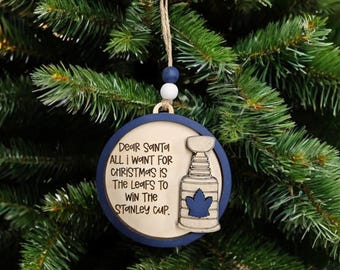 Pode incluir: Um enfeite de Natal de madeira azul e branco com o texto "Caro Papai Noel TUDO QUE EU QUERO PARA O NATAL É QUE OS LEAFS GANHEM A COPA STANLEY." e um gráfico de um troféu de hóquei.