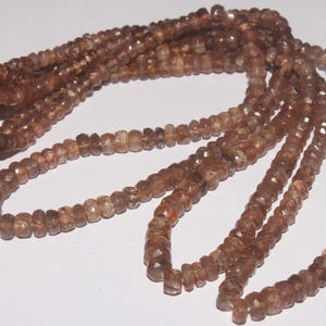 May include: Strands of brown gemstone beads. The beads are small, faceted, and have a warm, earthy tone. The strands are arranged in a flowing pattern, suggesting they are part of a necklace or bracelet. The background is a plain white surface.