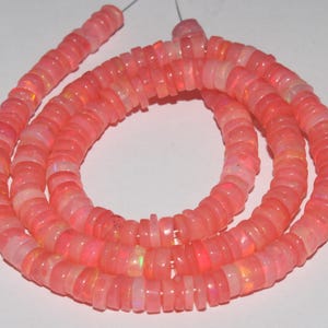 May include: A strand of circular, disc-shaped beads in a vibrant coral pink hue. Each bead features a smooth, polished surface with subtle, iridescent flashes of light reflecting across the material, strung together on a thin, clear filament.