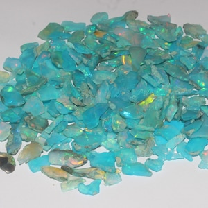 May include: A pile of raw, rough-cut opal gemstones. The opals are primarily a vibrant turquoise color, with flashes of iridescent color. The gemstones are irregularly shaped and vary in size, creating a natural, organic appearance.