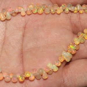 Peut inclure: Un collier de perles d'opale, composé de pierres en forme de goutte et de fleur. Les opales présentent une gamme de couleurs, dont le blanc, le jaune et l'orange, avec des reflets irisés. Le collier est monté sur un cordon fin et transparent et possède des pompons noirs.