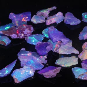 May include: A collection of raw opal gemstones in various shapes and sizes. The opals display a range of colors, including blue, purple, and green, with iridescent flashes. The gemstones are scattered on a dark surface.