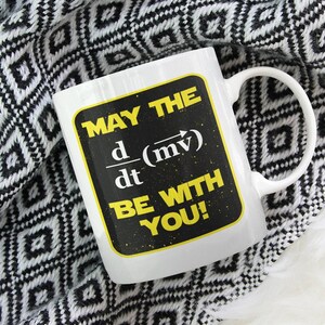 May include: White ceramic mug with a black and yellow design. The mug has the text "May the d(mv)/dt be with you!" printed on it.