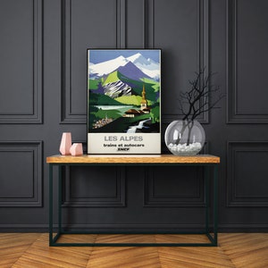 May include: A vintage travel poster advertising travel to the Alps by train and bus. The poster features a colorful illustration of a mountain range with a church and a waterfall. The text on the poster reads "Les Alpes trains et autocars SNCF".