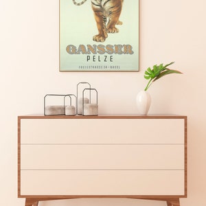May include: A framed vintage poster featuring a roaring tiger with the text "GANSSER PELZE" in brown letters. The poster is displayed above a modern, three-drawer wooden dresser with a white finish and angled legs. A white vase and decorative candle holders are on the dresser.