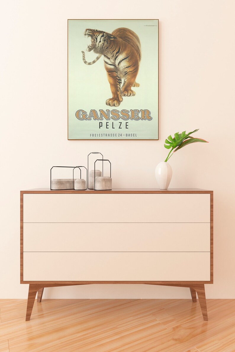May include: A framed vintage poster featuring a roaring tiger with the text "GANSSER PELZE" in brown letters. The poster is displayed above a modern, three-drawer wooden dresser with a white finish and angled legs. A white vase and decorative candle holders are on the dresser.