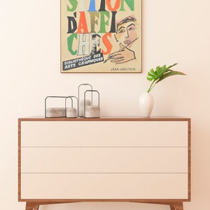 May include: A vintage poster with the text "EXPO.  SITION DAFFI CHESS" in colourful, bold letters. The poster features a drawing of a person's hand holding a paintbrush and a close-up of a person's face. The text "BIBLIOTHEQUE DES ARTS GRAPHIQUES" and "JEAN ARESTEIN" are at the bottom of the poster.