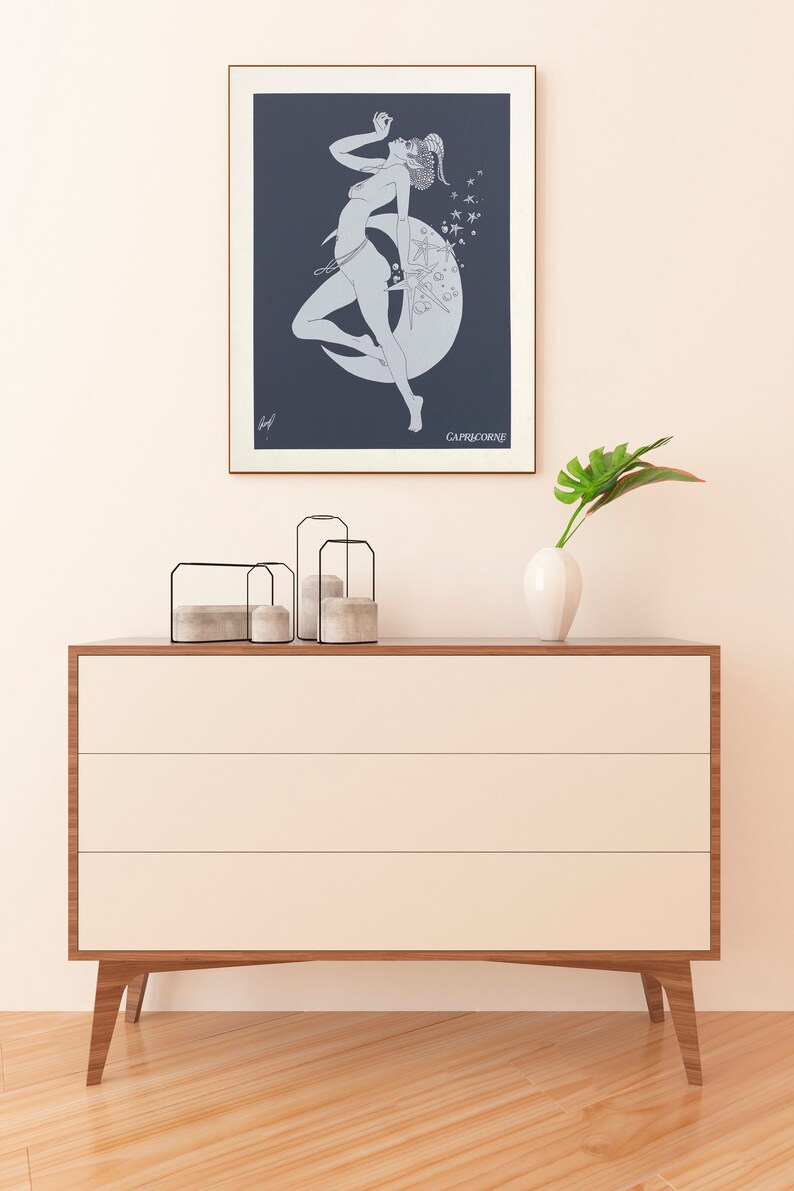 May include: A framed print of a line drawing of a woman standing on a crescent moon with stars in the background. The text "CAPRICORNE" is at the bottom of the print.
