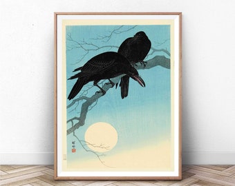 Puede incluir: Una impresión japonesa vintage en madera que representa dos cuervos negros posados en una rama desnuda contra un cielo azul con luna llena. La impresión está enmarcada en un marco de madera claro.