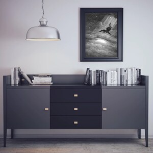 K&ouml;nnte beinhalten: Ein Schwarzwei&szlig;druck einer gefl&uuml;gelten Figur, die vom Himmel f&auml;llt, in Schwarz gerahmt und an einer wei&szlig;en Wand &uuml;ber einer dunkelgrauen Kommode mit drei Schubladen und zwei T&uuml;ren h&auml;ngend. Die Kommode ist mit einer Reihe von B&uuml;chern bedeckt.
