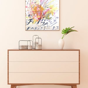 May include: A framed watercolor painting of a colorful abstract design with the text "Festival de Jazz Paris 25.tout. 1 Nov. 83". The painting is hanging on a white wall above a wooden dresser with three drawers.