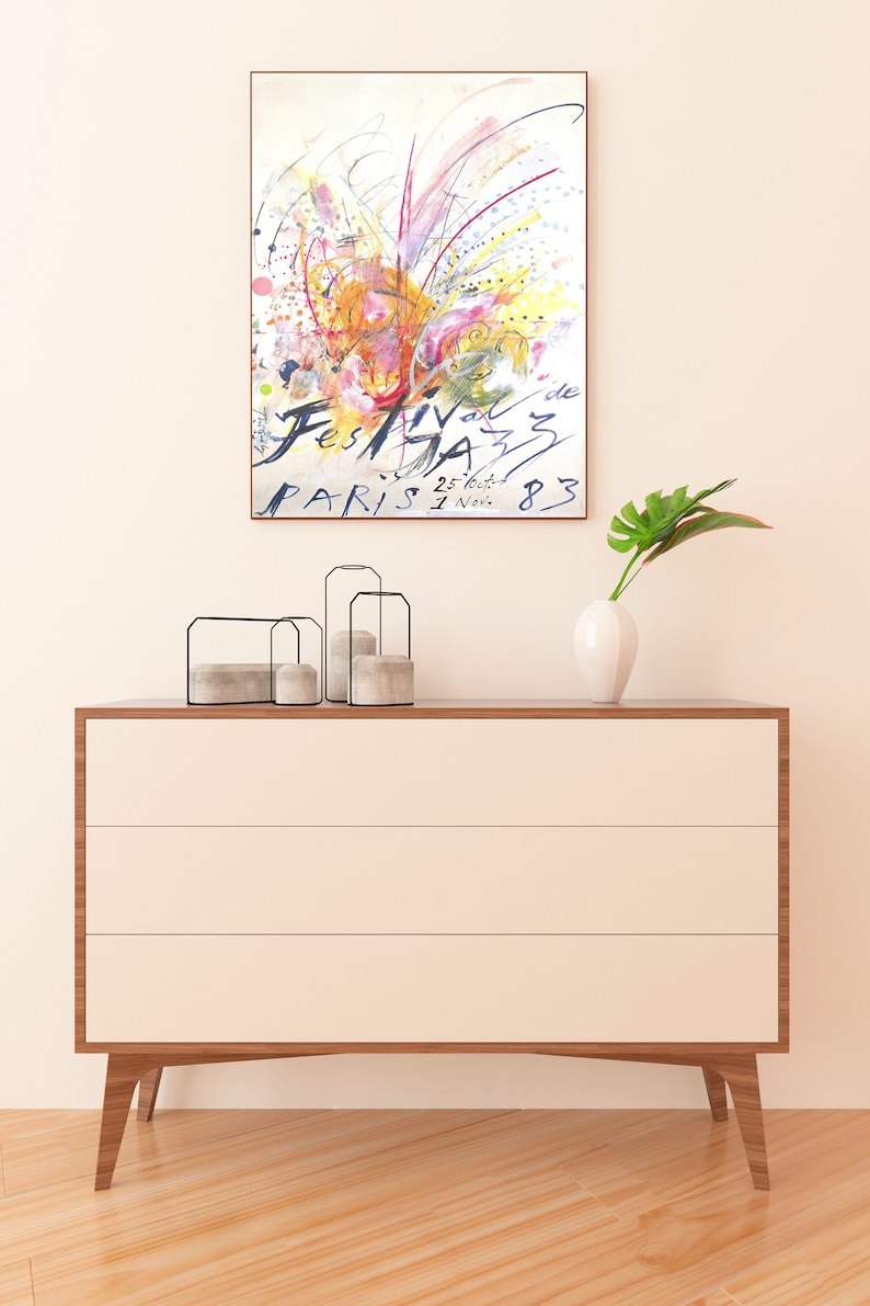 May include: A framed watercolor painting of a colorful abstract design with the text "Festival de Jazz Paris 25.tout. 1 Nov. 83". The painting is hanging on a white wall above a wooden dresser with three drawers.