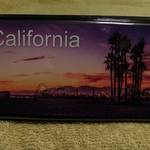 May include: A black rectangular box with a colorful image of a sunset over a beach with a Ferris wheel and palm trees. The text "California" is printed in white at the top of the image.