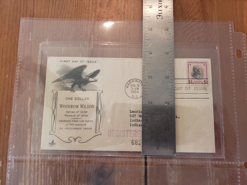 May include: A white envelope with a "First Day of Issue" stamp featuring a bald eagle and the text "One Dollar Woodrow Wilson Series of 1938 Reissue of 1954 Engraved from Flat Plates 400-Subjects On Presummed Paper". The envelope also has a "First Day of Issue" stamp with a portrait of Woodrow Wilson and the text "$1. $1".
