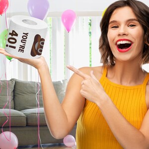 K&ouml;nnte beinhalten: Eine wei&szlig;e Toilettenpapierrolle mit dem Text "You're 40!" und einem Cartoon-Kack-Emoji. Die Rolle wird von einer Hand vor einem verschwommenen Hintergrund eines Wohnzimmers mit Ballons gehalten.