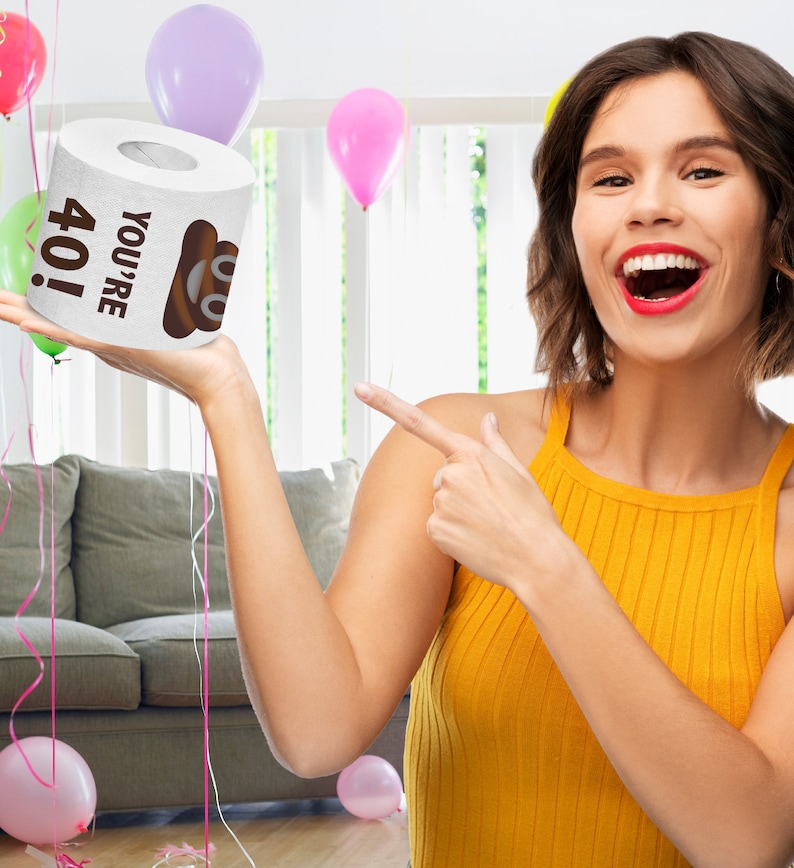 K&ouml;nnte beinhalten: Eine wei&szlig;e Toilettenpapierrolle mit dem Text "You're 40!" und einem Cartoon-Kack-Emoji. Die Rolle wird von einer Hand vor einem verschwommenen Hintergrund eines Wohnzimmers mit Ballons gehalten.