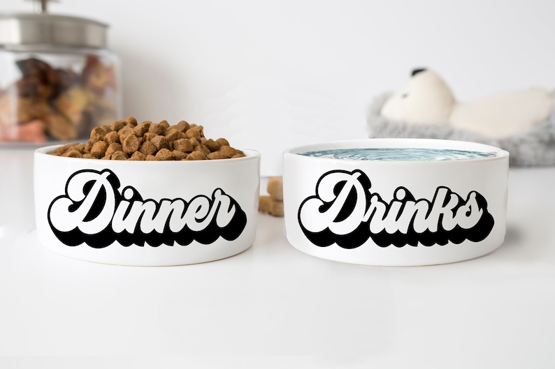 Op de afbeelding: Twee witte keramische dierenbakken met zwarte tekst. De bak links zegt "Dinner" en de bak rechts zegt "Drinks".