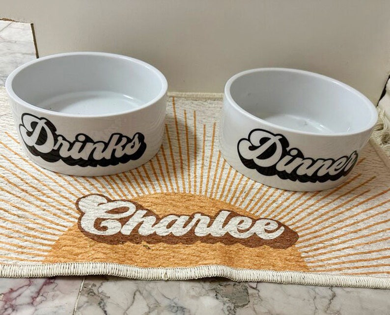 Op de afbeelding: Twee witte keramische huisdierkommen met zwarte letters. De kom links zegt "Drinks" en de kom rechts zegt "Dinner". Een bruin en wit kleed met de naam "Charlee" in bruine letters ligt onder de kommen.