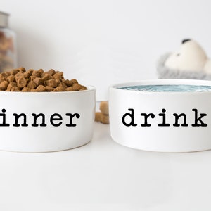 May include: Two white ceramic pet bowls, one filled with dry dog food and labeled "dinner", the other filled with water and labeled "drinks".