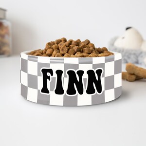 May include: A white and gray checkered dog bowl filled with brown kibble. The bowl has the name "FINN" in bold, black letters. A jar of treats and a stuffed animal are in the background.