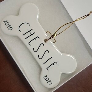 May include: White ceramic bone-shaped ornament with the name "Chessie" and the years "2010" and "2021" printed on it. The ornament has a gold string attached for hanging.